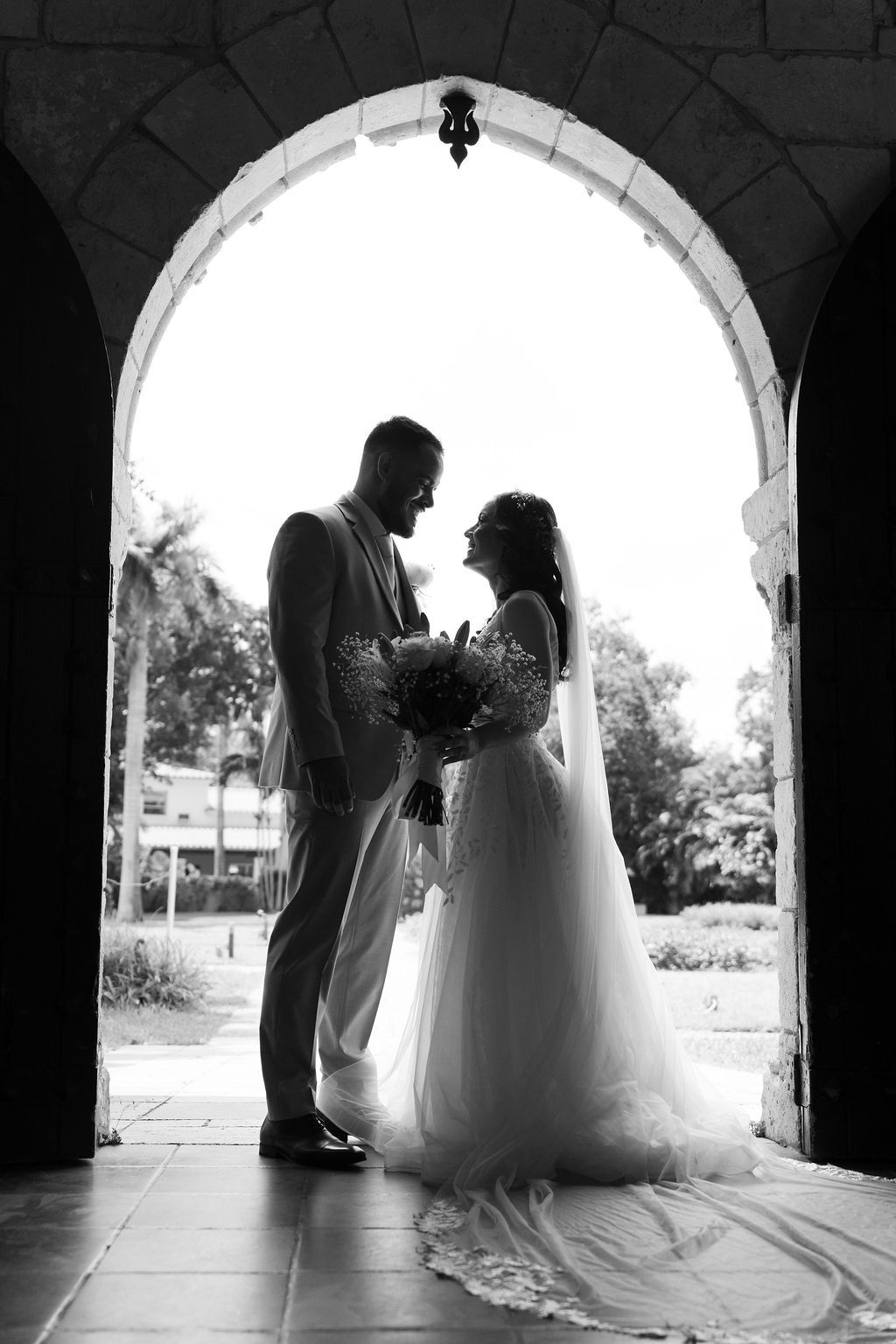 Bride and groom smiling beneath a historic stone archway in a timeless black and white wedding portrait, photographed by Miami wedding photo