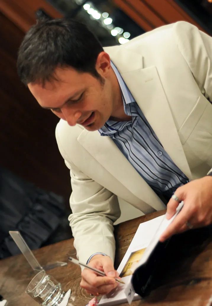 Man in white blazer signing a book at a table indoors.