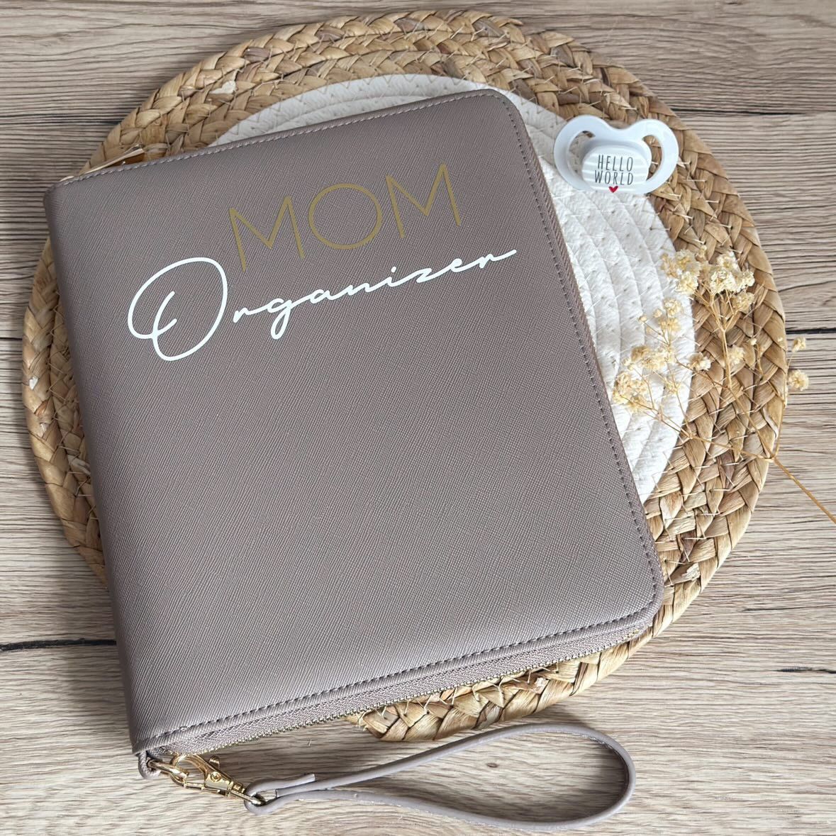 Gray faux leather MOM Organizer zipper binder with gold text, wrist strap, displayed on woven placemat with pacifier.