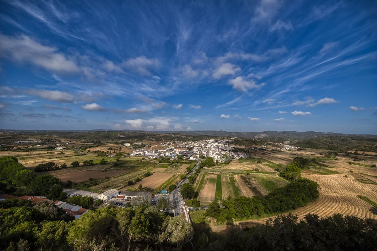 Aerial panoramic view of a small Mediterranean village surrounded by green farmland fields under a dramatic blue sky.