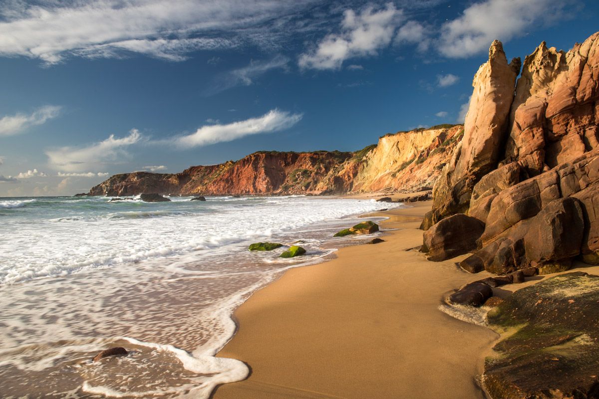 Golden sandy beach with red sandstone cliffs and rock formations under a blue sky with waves crashing ashore.