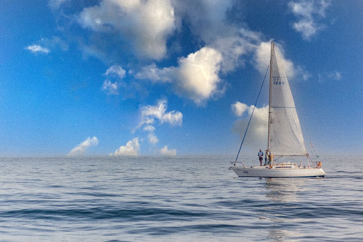 A white sailboat glides across calm blue ocean waters under a partly cloudy sky on a sunny day.