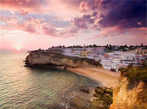 Stunning sunset over a cliffside Mediterranean village with white buildings, golden beach, and dramatic pink and purple sky.