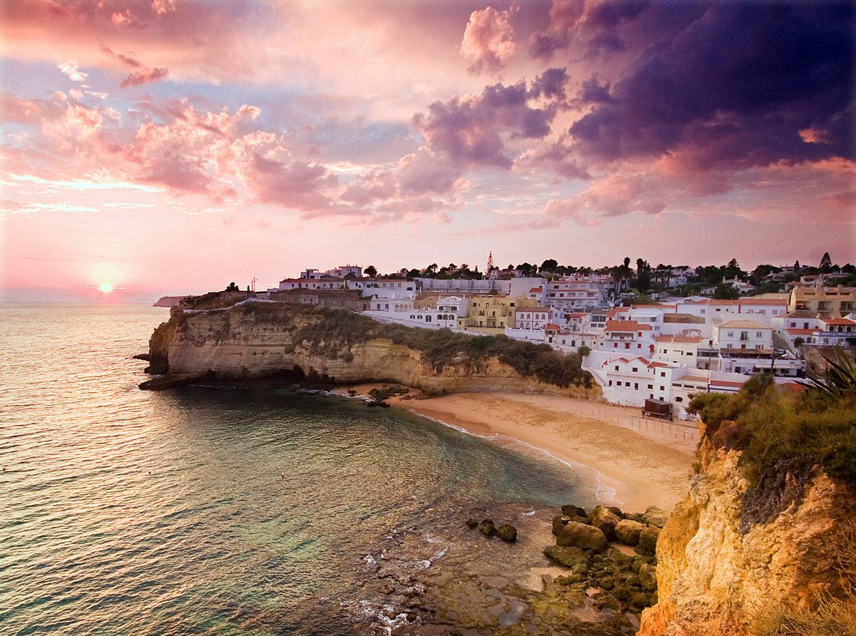 Stunning sunset over a cliffside Mediterranean village with white buildings, golden beach, and dramatic pink and purple sky.
