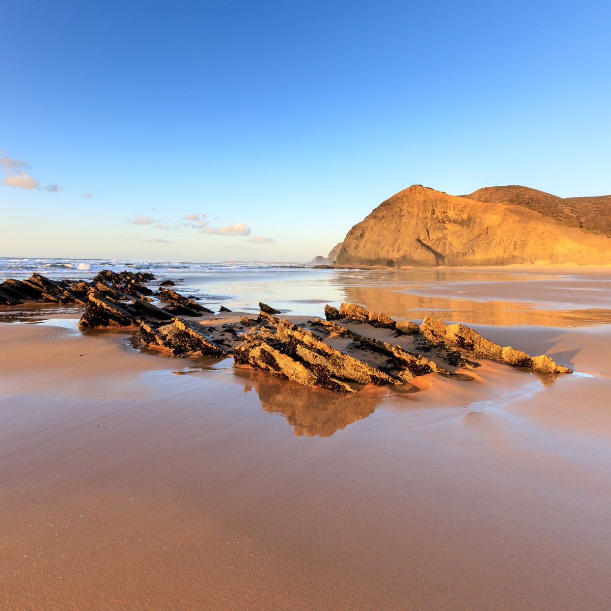 Golden hour sunlight illuminates rocky formations on a sandy beach with calm ocean waves and a dramatic cliff in the background.
