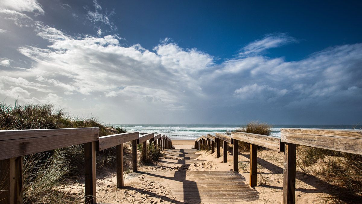 Wooden boardwalk path leading through sandy dunes to a sunny beach with ocean waves and dramatic cloudy sky.