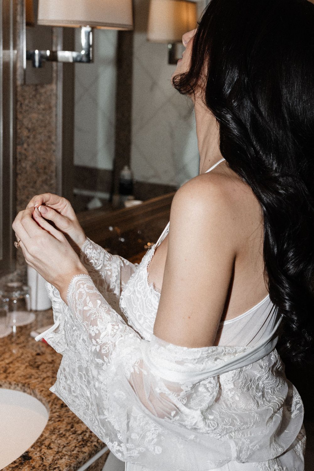 Bride with dark hair in lace robe adjusting jewelry at bathroom vanity mirror on wedding day.