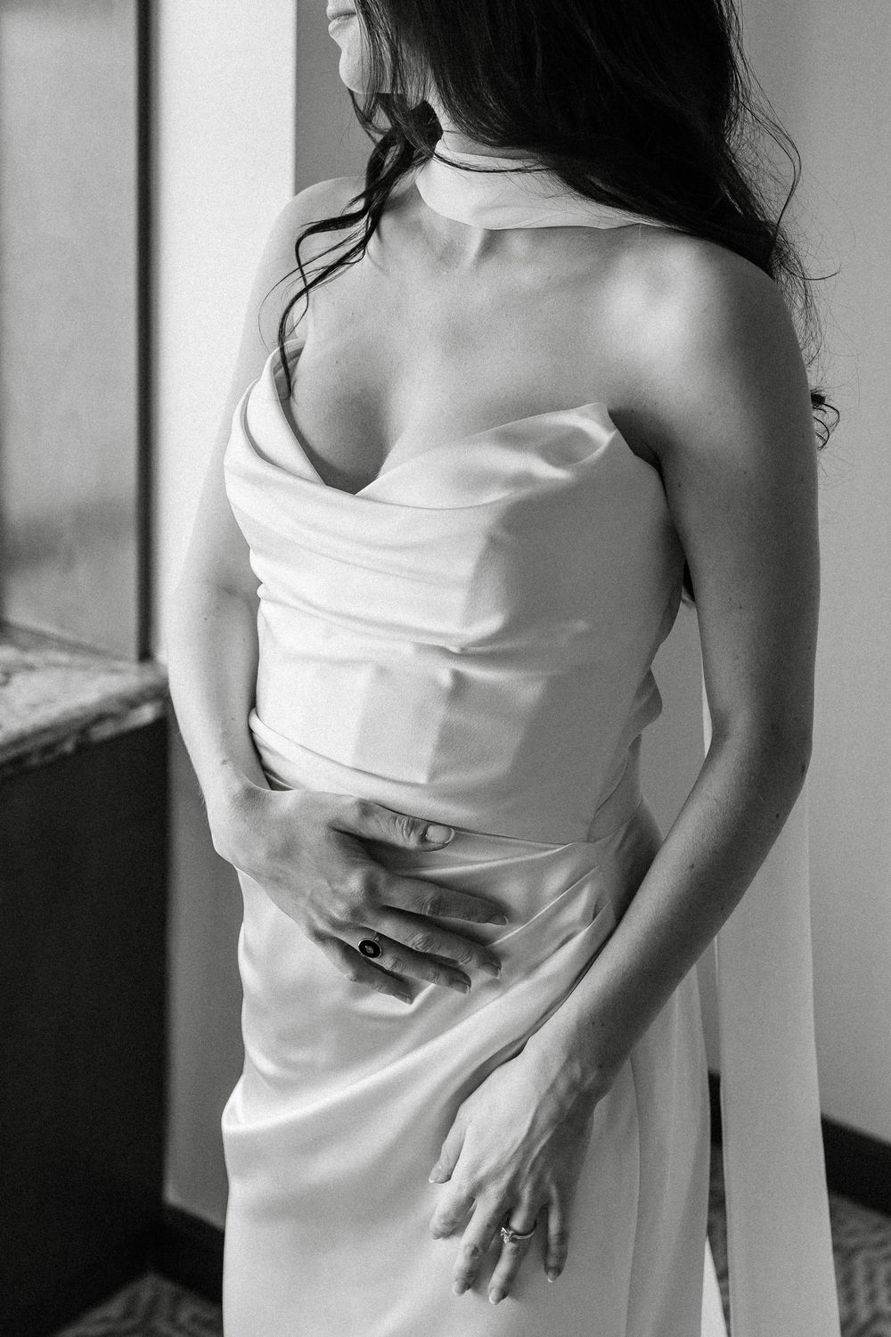 Black and white photo of a bride in a satin halter wedding gown, hands resting on her waist near a window.