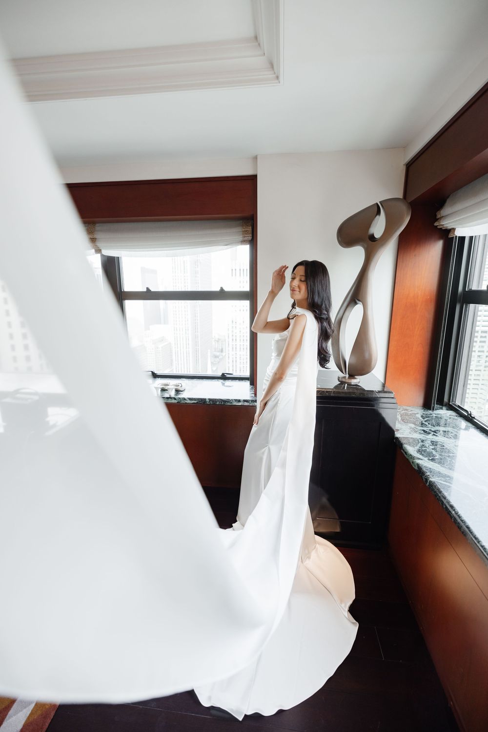 Bride in elegant white gown with long train stands by window in luxury hotel room, hand raised gracefully.