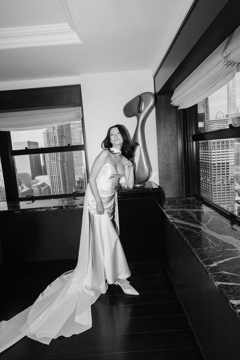 Black and white photo of a bride in a white gown posing elegantly in a high-rise hotel room with city skyline views.