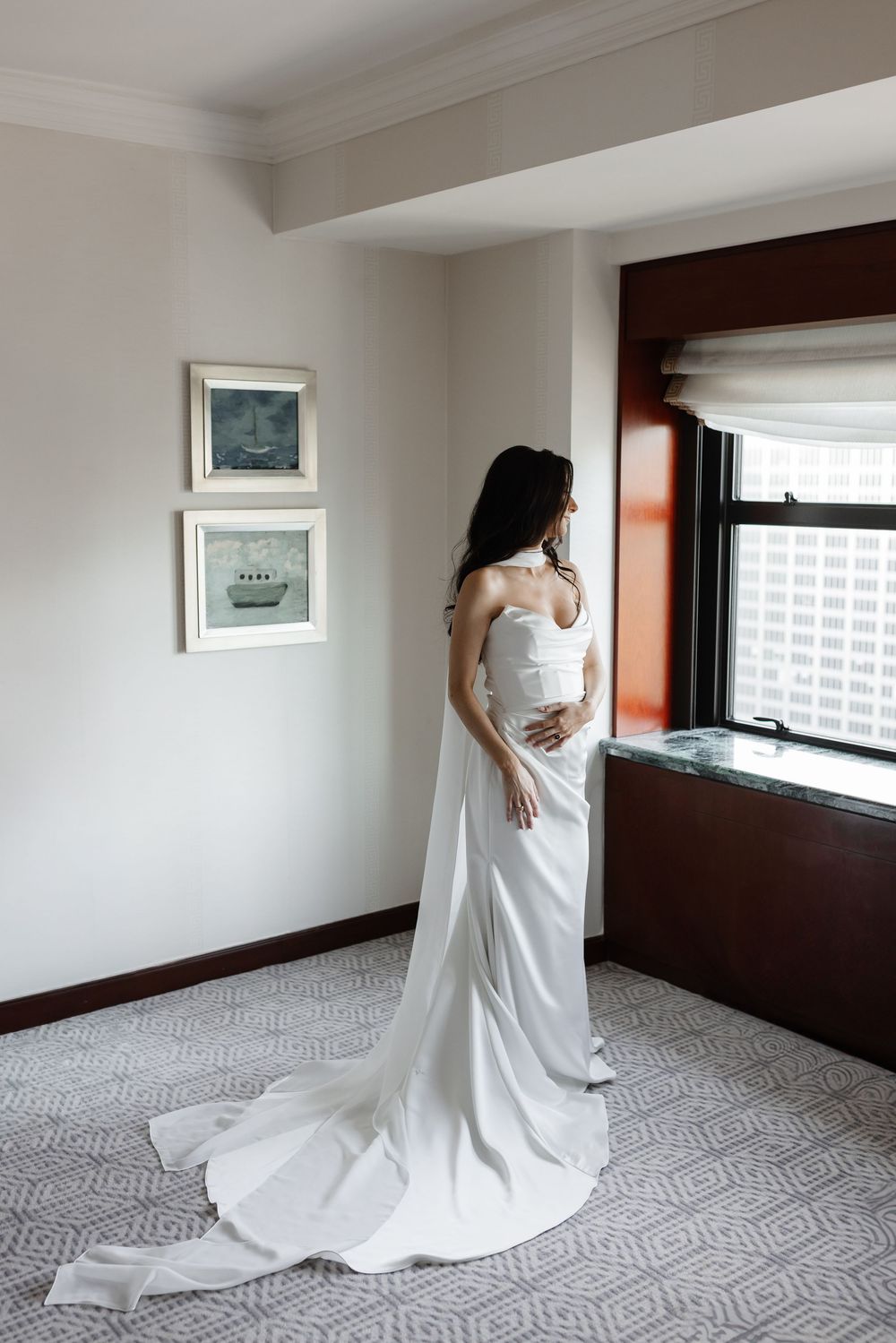 Bride in elegant white satin wedding gown with long train standing by hotel room window on wedding day.