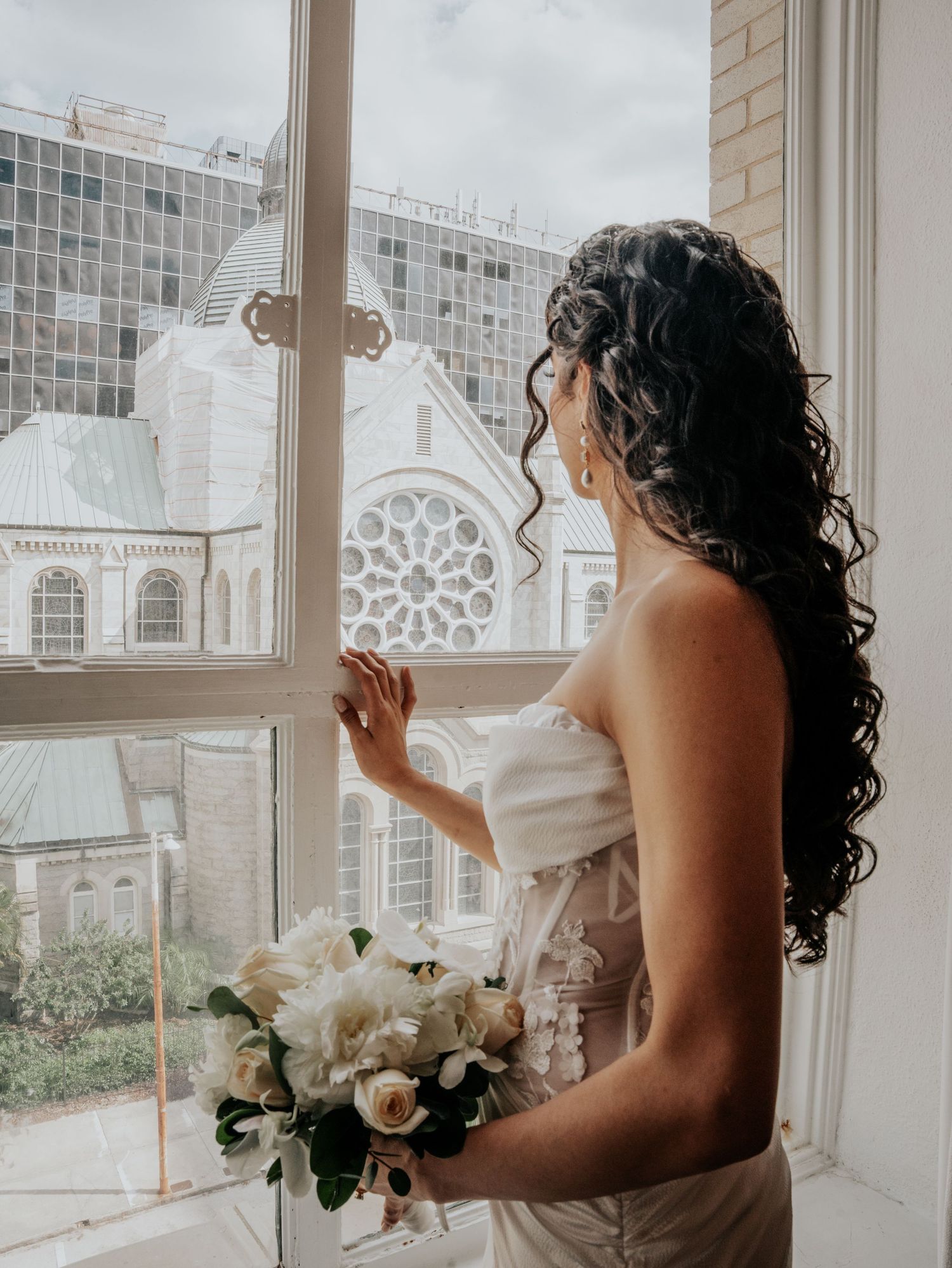 Bride with curly hair holding white bouquet gazing out window at historic church on wedding day.
