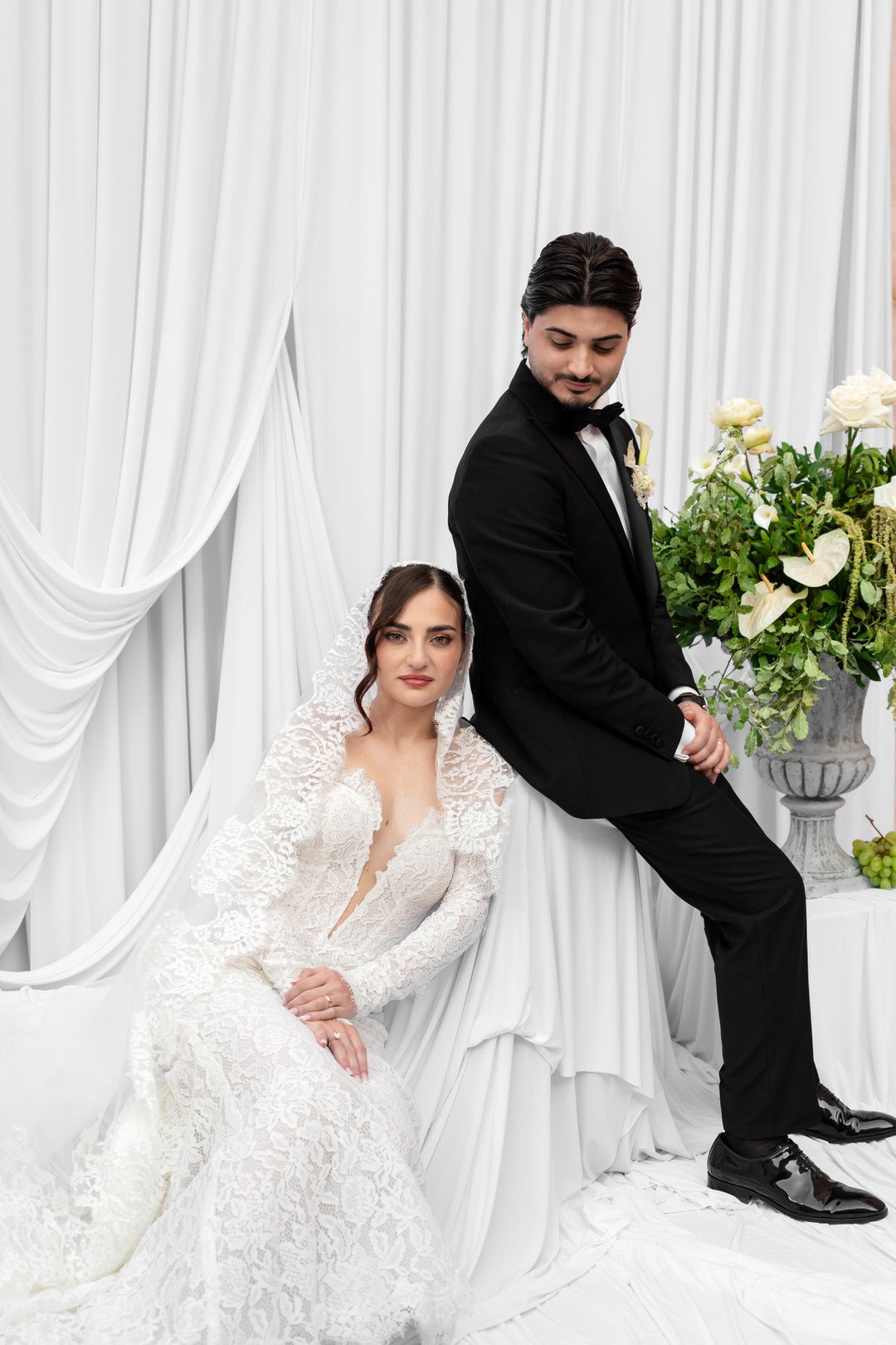 Elegant bride in white lace gown sits as groom in black tuxedo stands beside floral arrangement against white drape backdrop.
