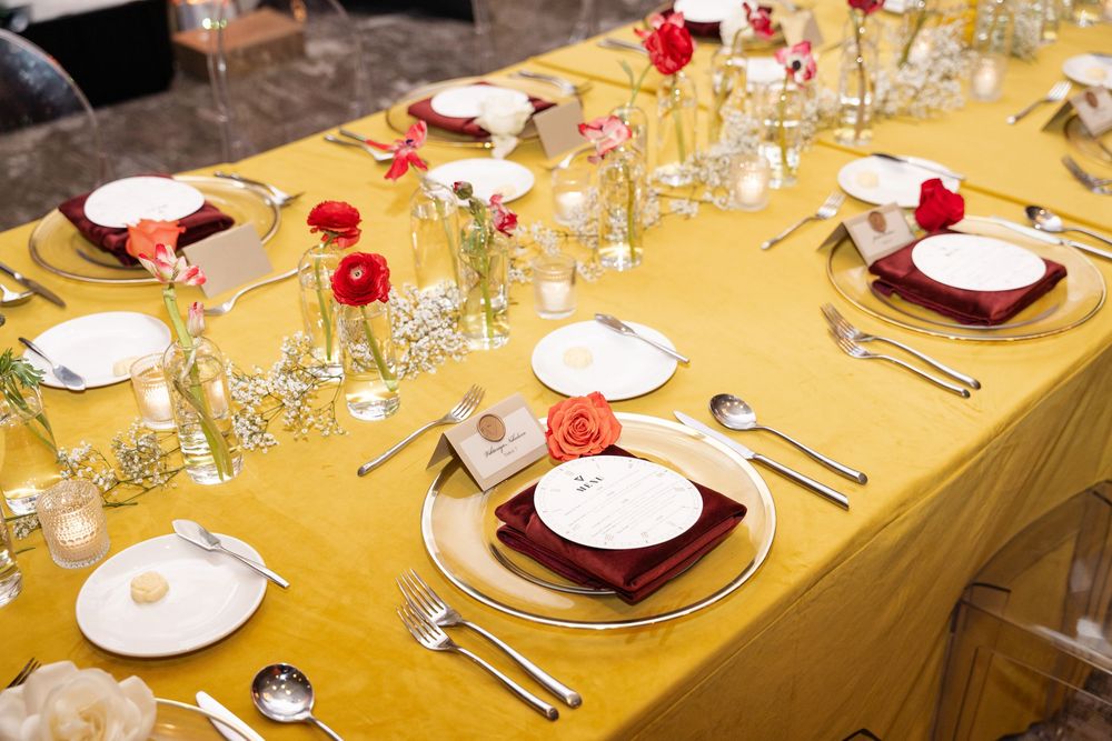Elegant wedding table setting with gold tablecloth, white plates, burgundy napkins, and red floral centerpieces.