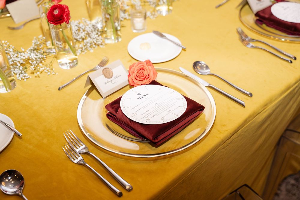 Elegant wedding table setting with gold tablecloth, burgundy napkin, circular menu card, and coral rose centerpiece.