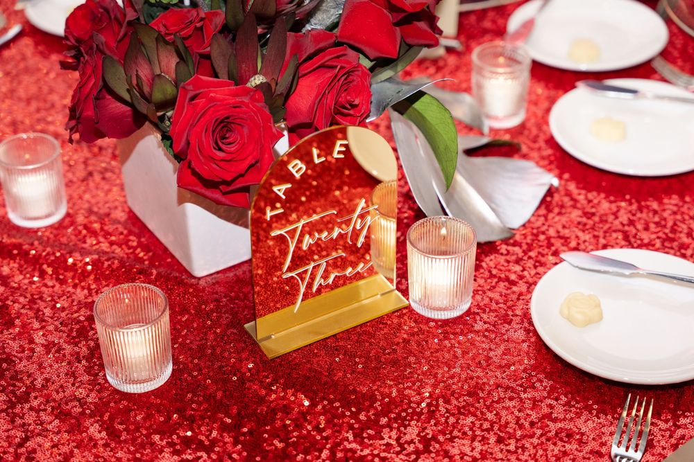 Red sequin tablecloth with gold table number sign, red roses centerpiece, and glowing candles at a wedding reception.