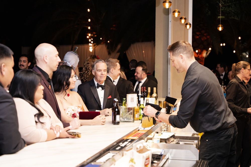 Guests gather at an outdoor bar during an evening event, with a bartender serving drinks under warm string lights.
