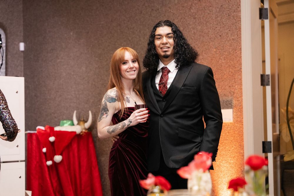 Couple posing at formal event, woman in burgundy velvet gown with tattoos, man in black suit with red tie.