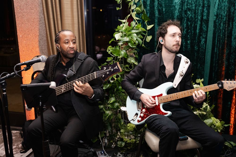 Two musicians playing electric guitars at an indoor event, dressed in black suits with green foliage backdrop.