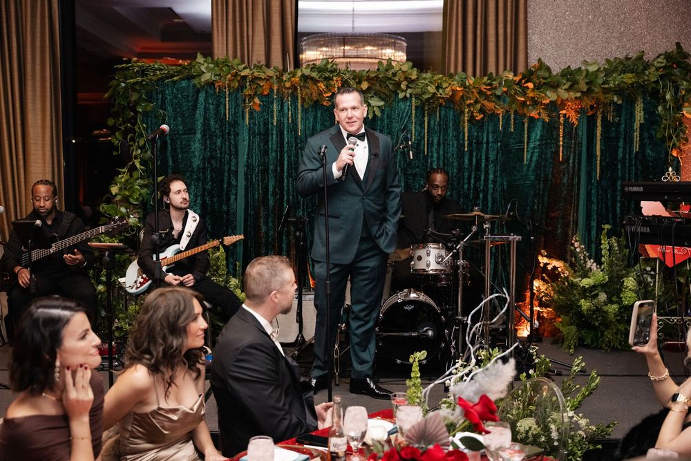 Man in tuxedo gives speech at elegant holiday wedding reception with live band and lush greenery decor.