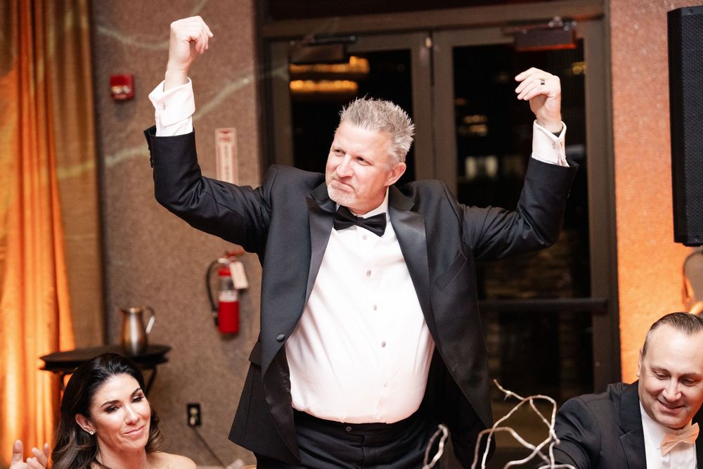 Man in tuxedo celebrating with arms raised at a formal wedding reception event.