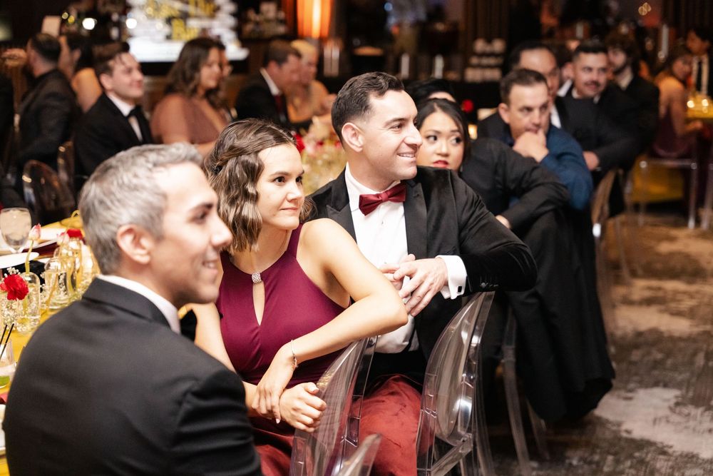 Elegantly dressed guests seated at a formal event, smiling and socializing in a warmly lit venue.