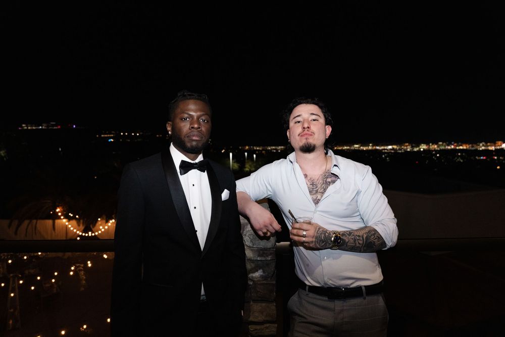 Two men pose outdoors at night with city lights in the background, one in a tuxedo and one in a white shirt.