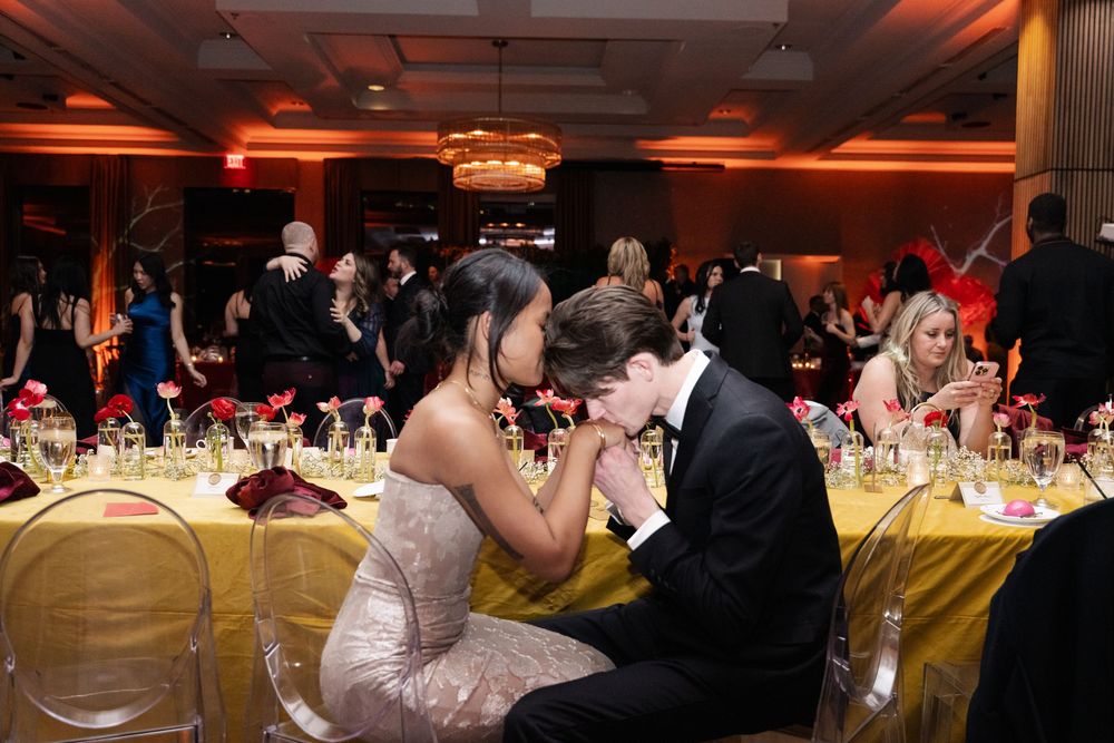 A couple shares a romantic moment at a formal gala, the young man kissing his date's hand at a elegantly set banquet table.