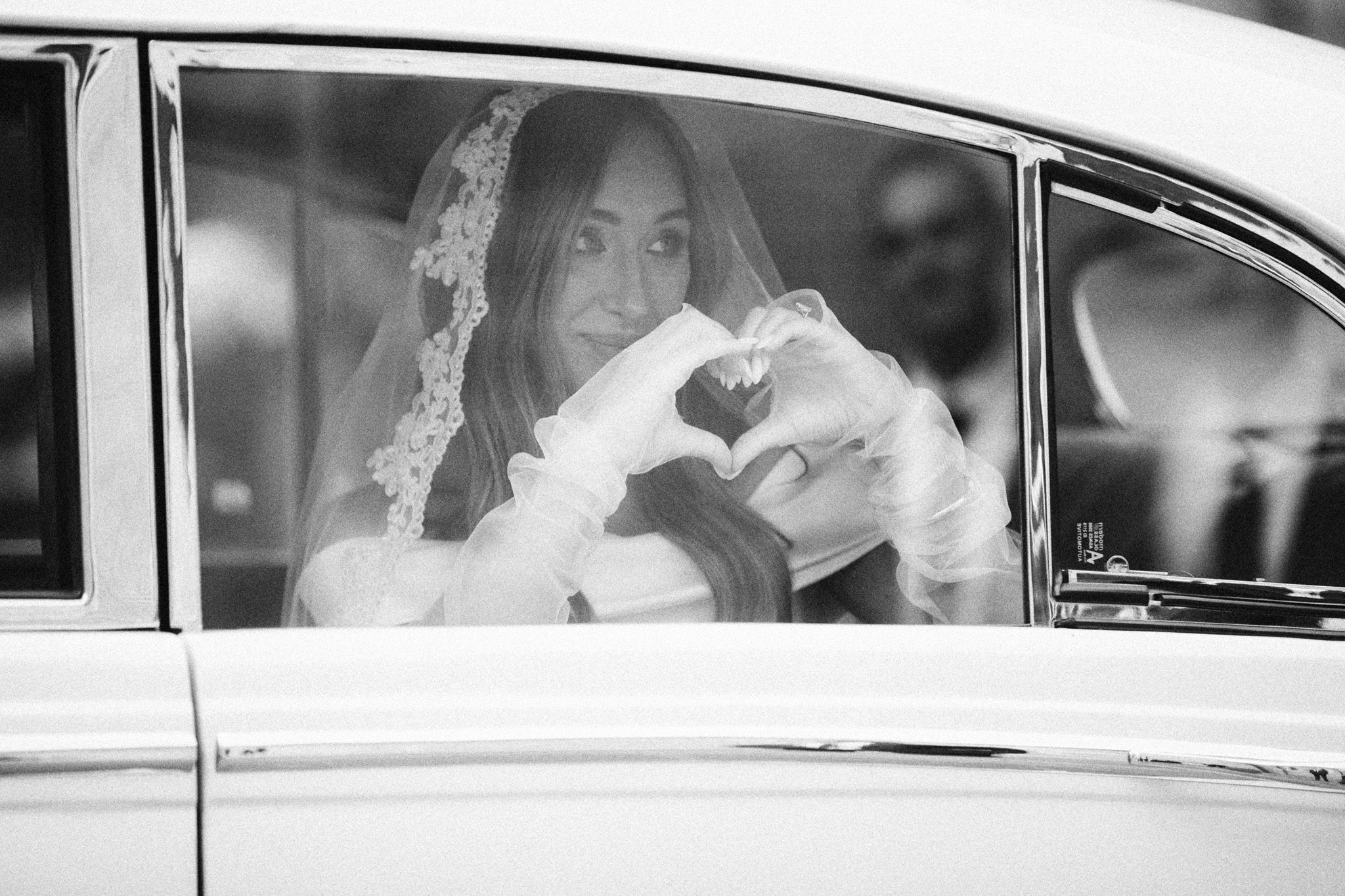 Bride in veil makes heart shape with hands through classic car window in elegant black and white wedding photo.