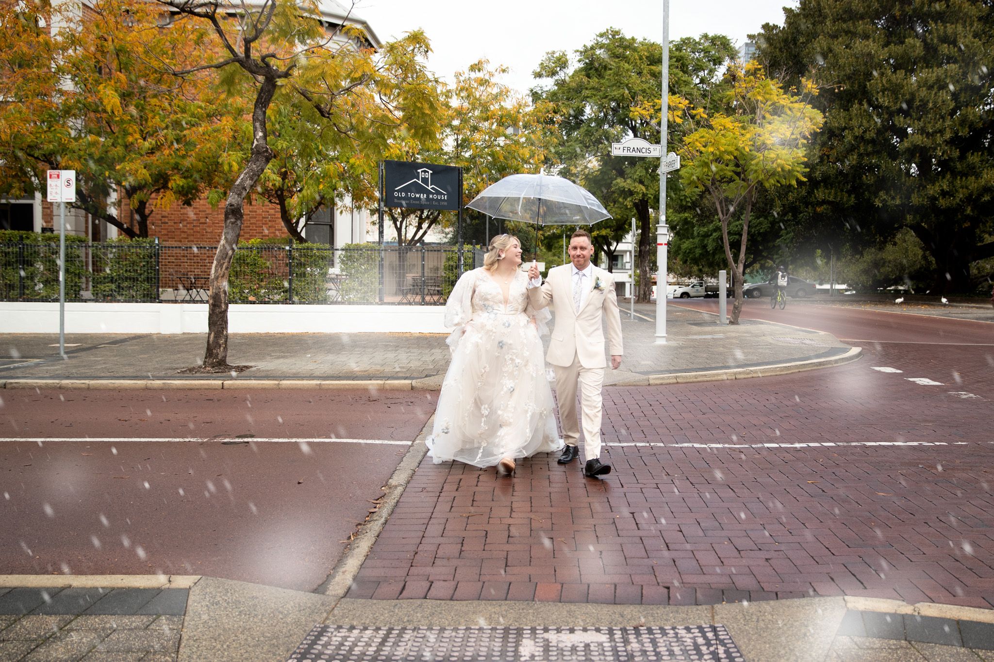 Bride and groom walking under umbrella in rain on brick crosswalk, surrounded by autumn trees.