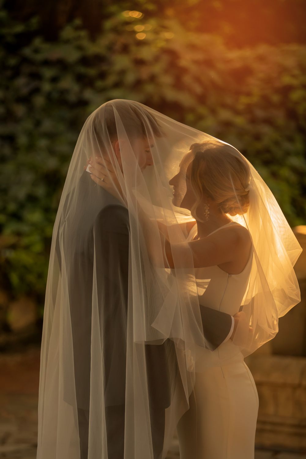 Bride and groom share romantic moment under wedding veil at golden hour, glowing sunlight illuminating outdoor ceremony.