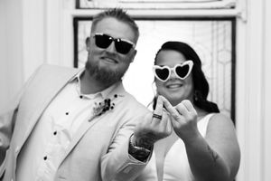 Black and white photo of a smiling couple in sunglasses showing off wedding rings, woman wearing heart-shaped frames.