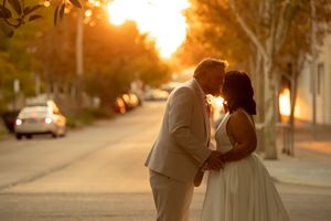 Couple sharing a romantic kiss at sunset on a tree-lined street, bride in white dress and groom in tan suit.