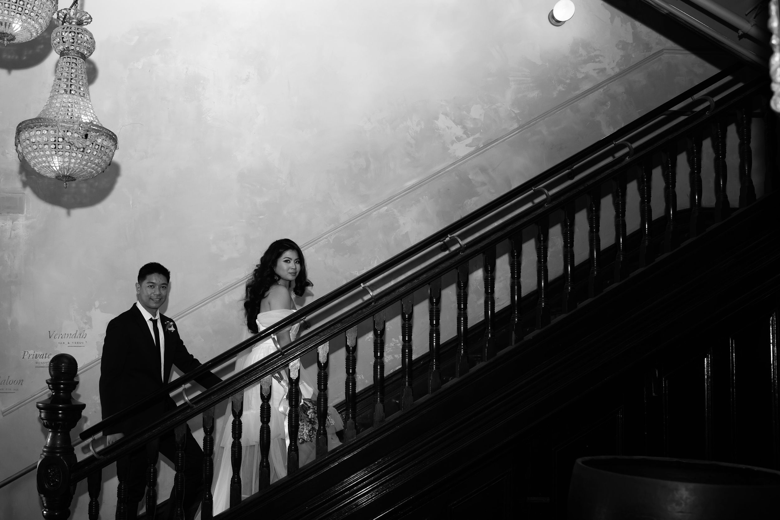 Black and white photo of a couple in formal attire descending an elegant staircase with a crystal chandelier.