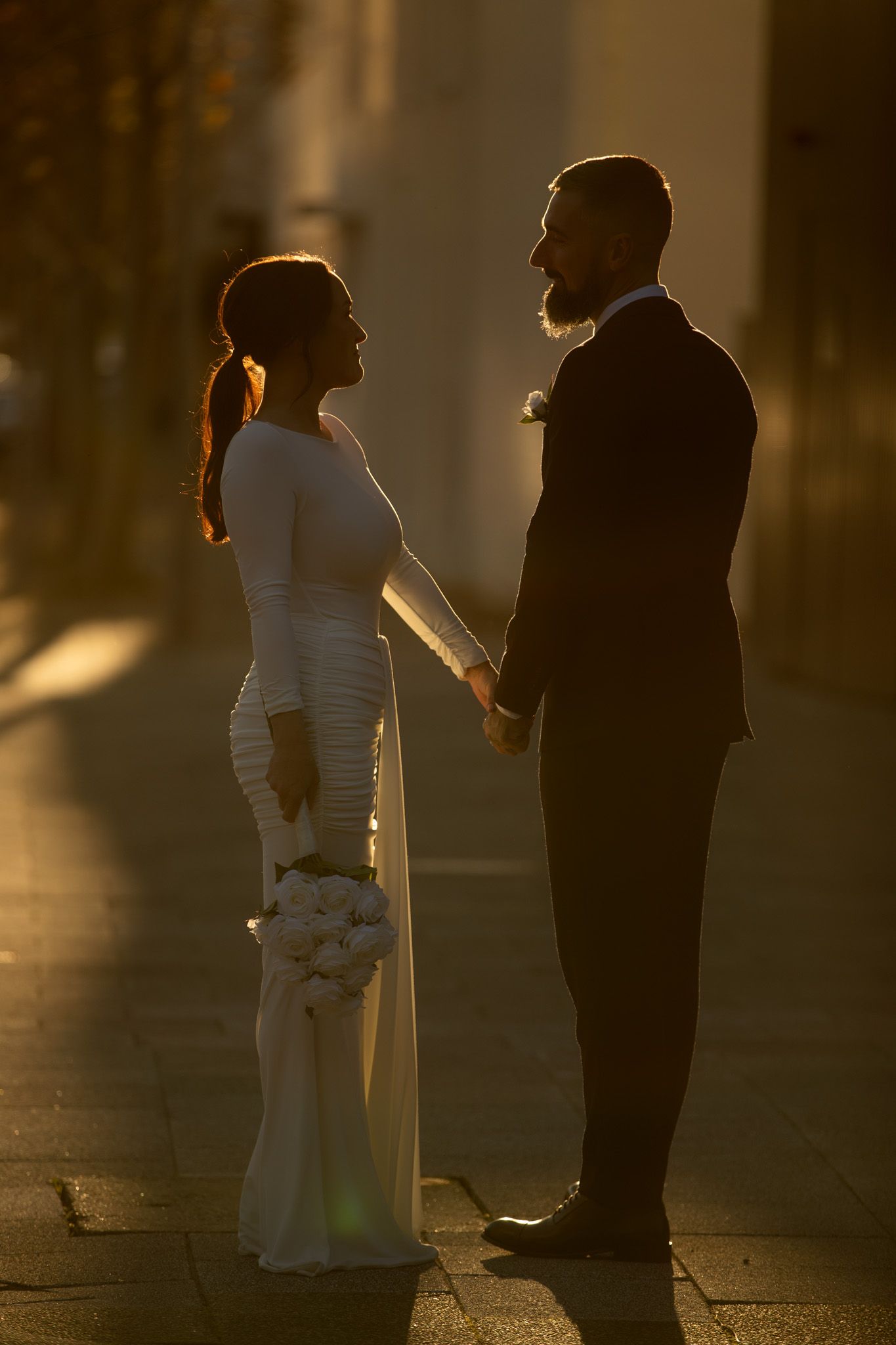 Bride and groom holding hands in golden sunset light, silhouetted on cobblestone street, bride in white gown holding bouquet.