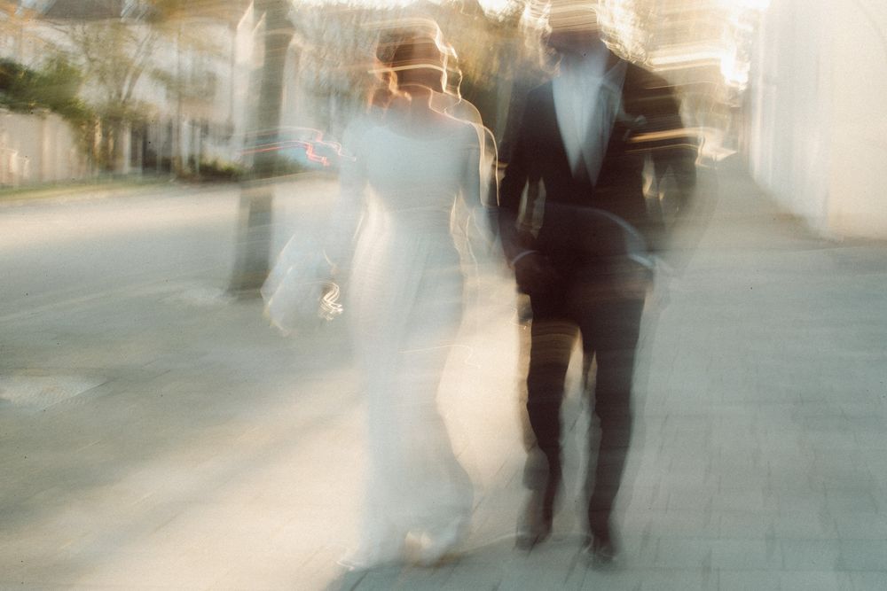 Blurred motion photo of bride in white gown and groom in black tuxedo walking together outdoors.