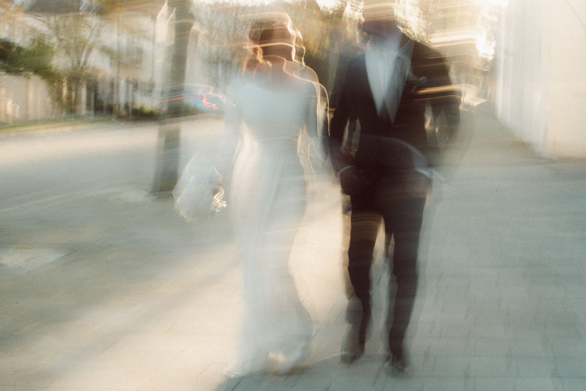 Blurred motion photo of bride in white gown and groom in black tuxedo walking together outdoors.