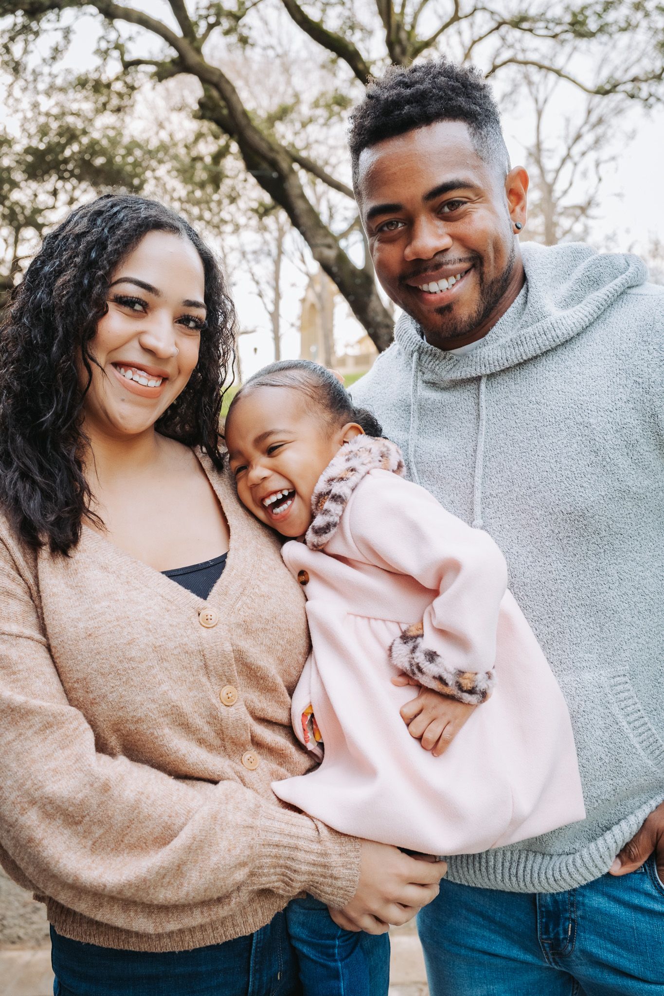 A happy Black family with a laughing toddler in pink poses outdoors near trees for a portrait.