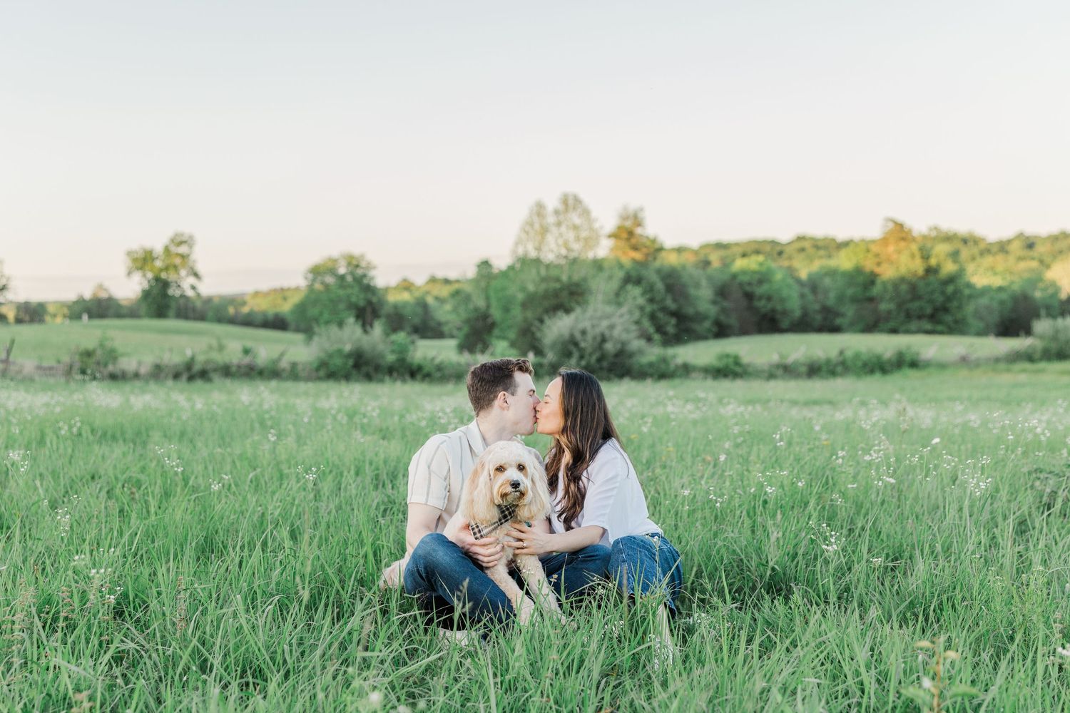 Couple sitting in green field kissing with golden doodle dog at golden hour during engagement photo session.
