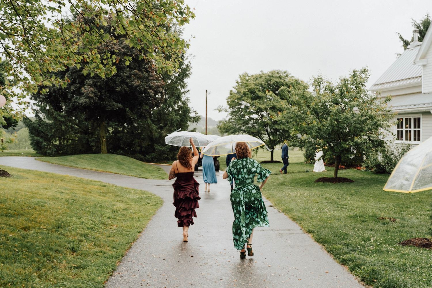 Two women in colorful dresses walk with umbrellas on a rainy garden path near a white building.
