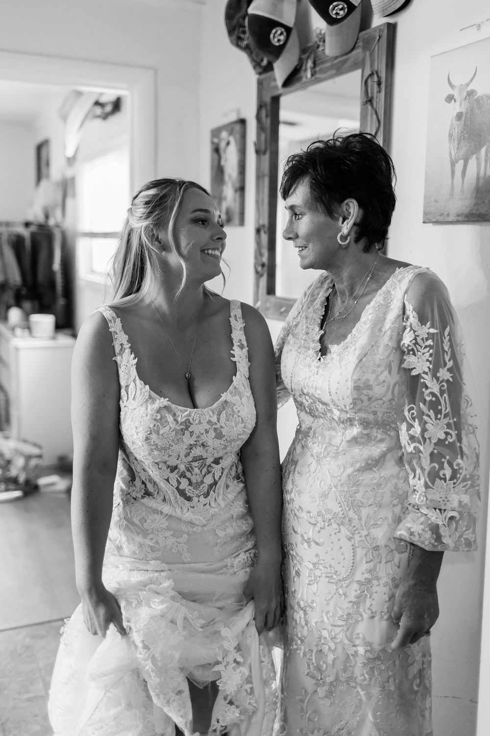 Two women in lace wedding dresses smiling at each other in a black and white bridal getting ready photo.