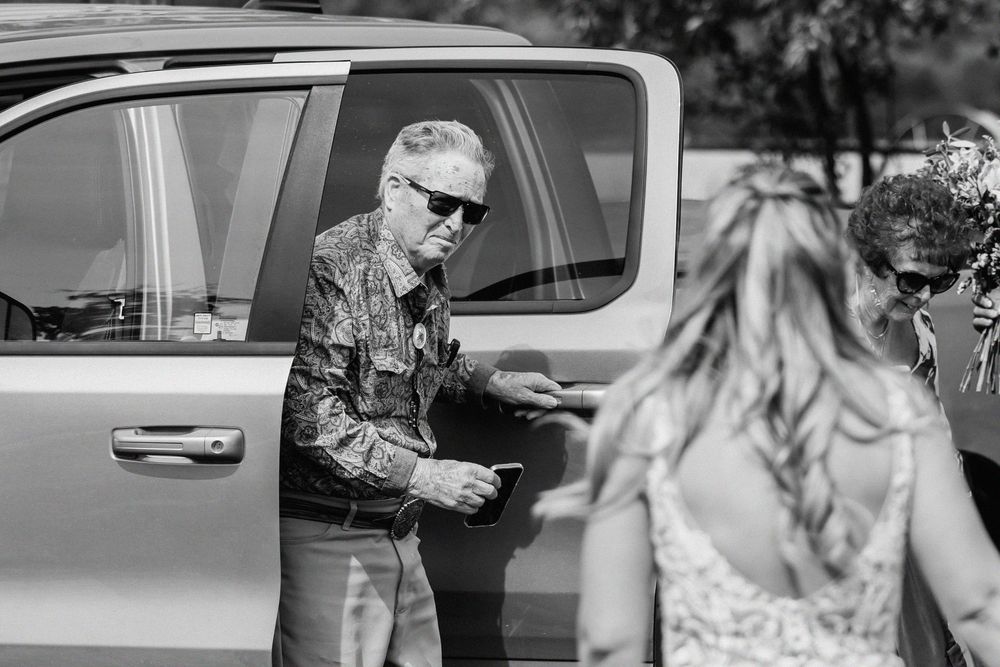 Black and white photo of a man in sunglasses opening a truck door for a bride at a wedding.