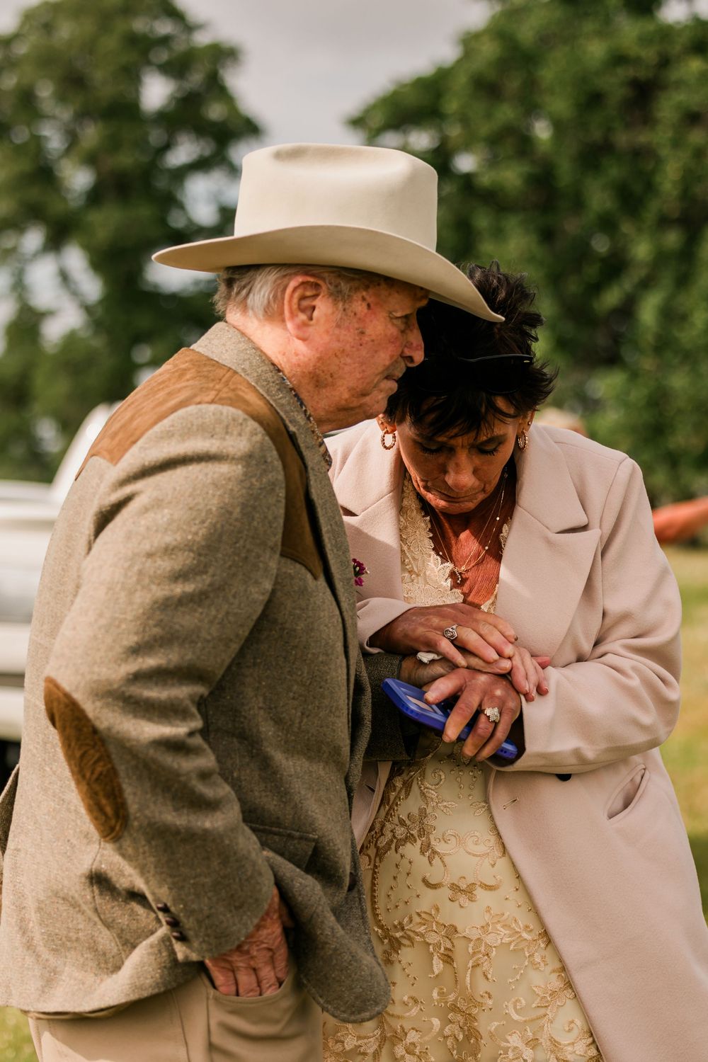 Elderly couple in formal attire examining a phone outdoors, man wearing cowboy hat and tweed jacket.