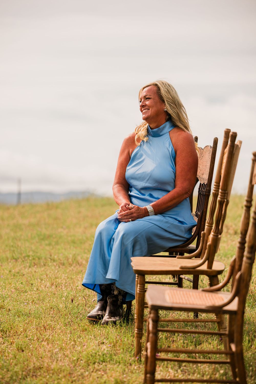 Woman in blue sleeveless dress sitting on wooden chair outdoors on green grass, smiling joyfully.