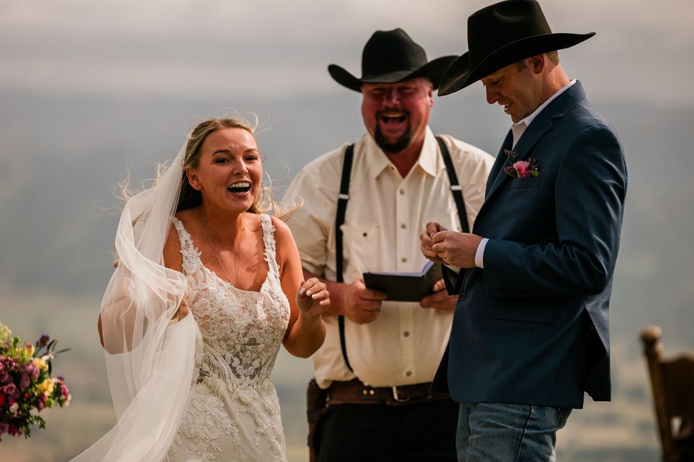 Bride and groom laughing joyfully during outdoor wedding ceremony with officiant in cowboy hat.