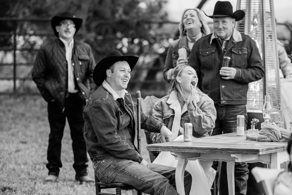 Black and white photo of laughing friends in cowboy hats gathered around an outdoor table at a country event.