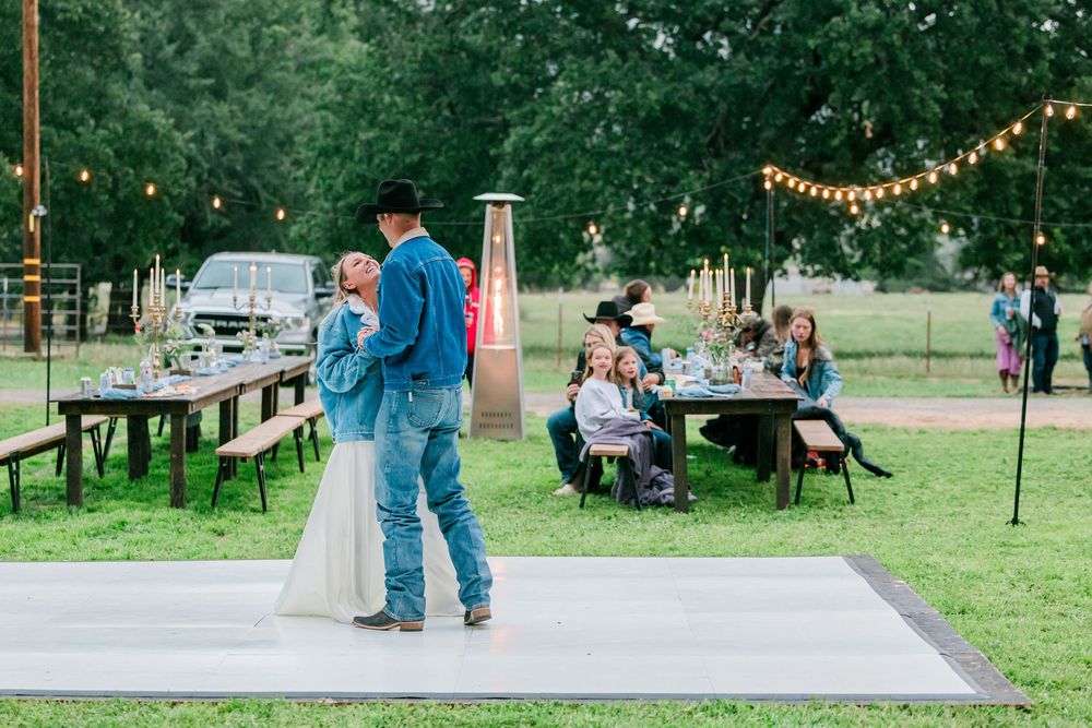 Outdoor wedding reception with couple dancing on white floor, string lights, and guests at rustic wooden tables in a green field.