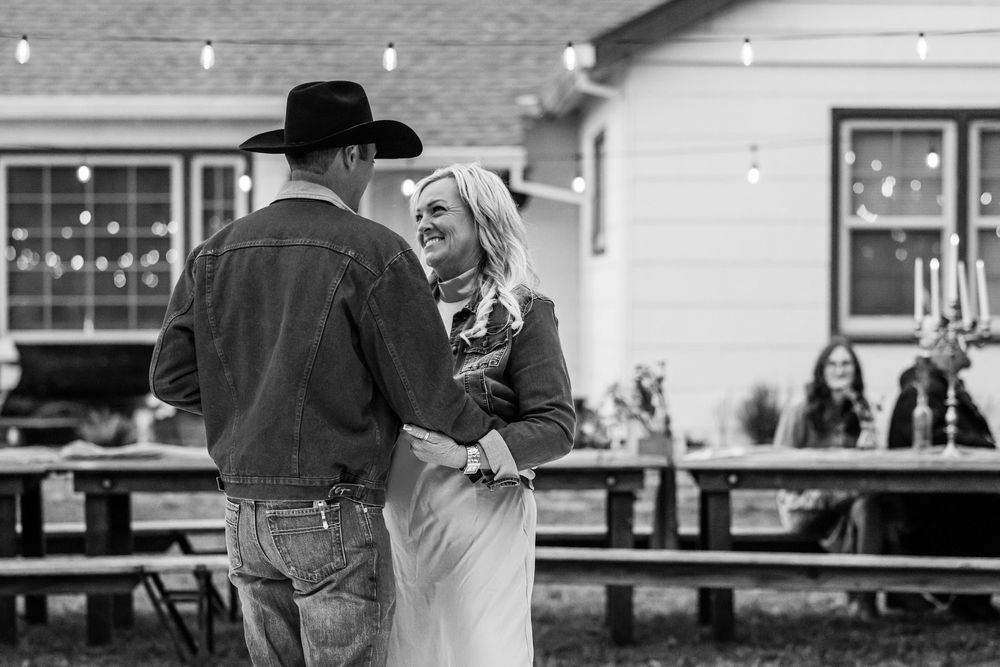 Couple sharing a joyful moment at an outdoor rustic wedding reception with string lights and guests in background.