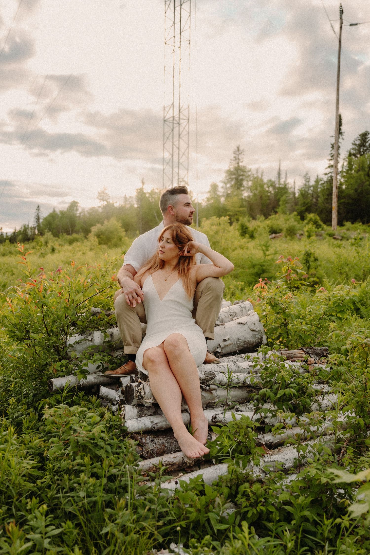 Couple sitting on birch logs in a wildflower meadow at golden hour, woman in white dress leaning against man in white shirt.