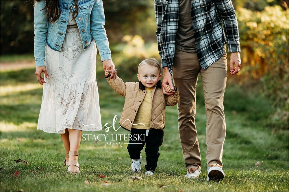 A young family walks hand-in-hand outdoors, parents guiding their toddler through a lush green park in autumn.